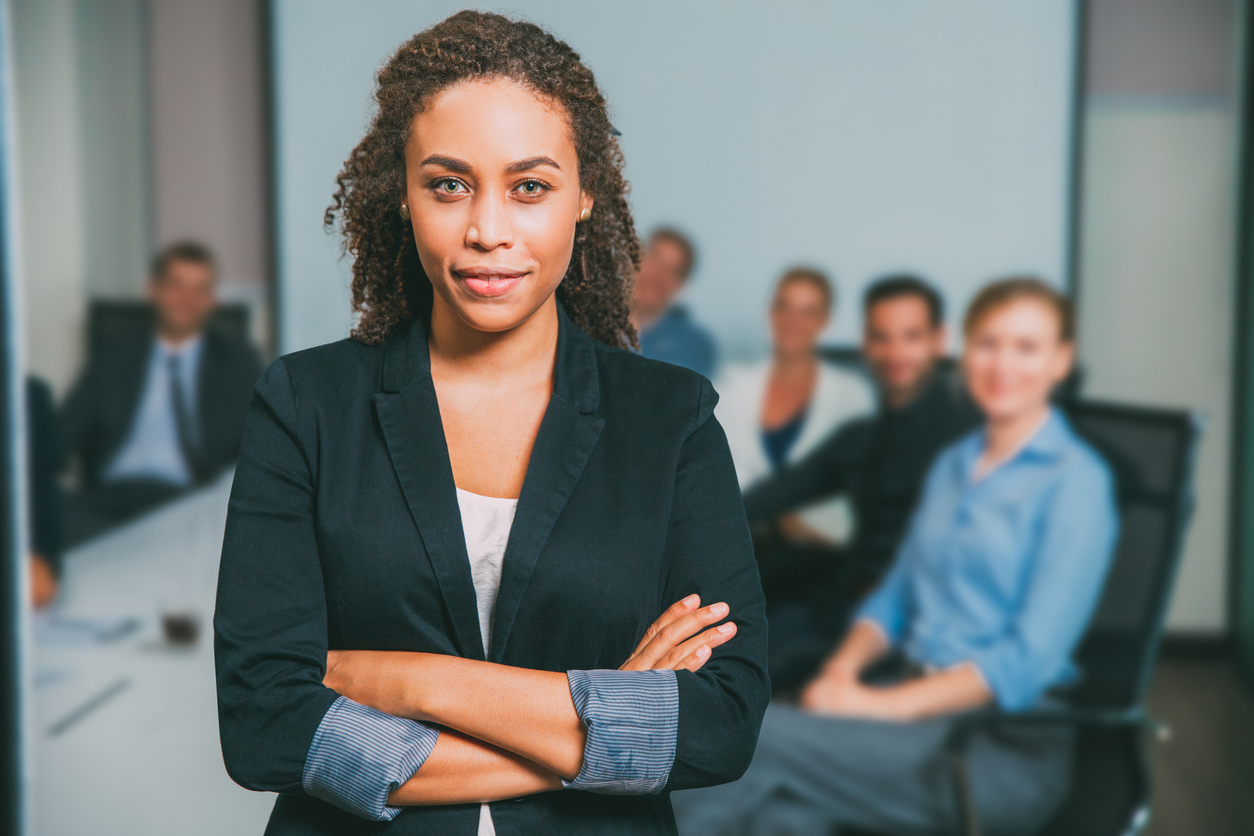 Mulher negra CFO se posiciona de braços cruzados sorrindo em frente a uma equipe de colaboradores.