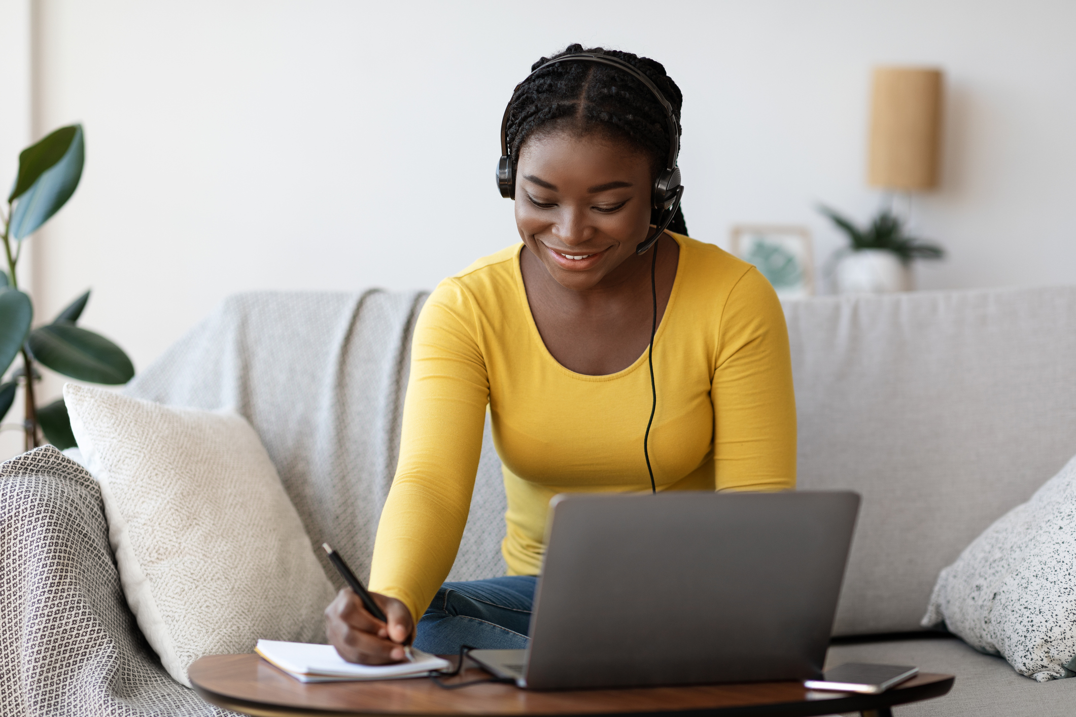 Mulher negra veste uma camiseta amarela sentada no sofá enquanto mexe num computador com um headphone e escreve com uma caneta em um papel anotações.