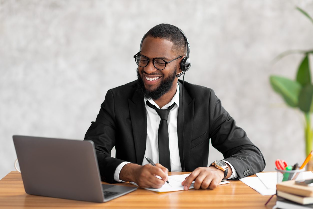 Homem negro estudo em frente a um computador, fazendo anotações e sorrindo.